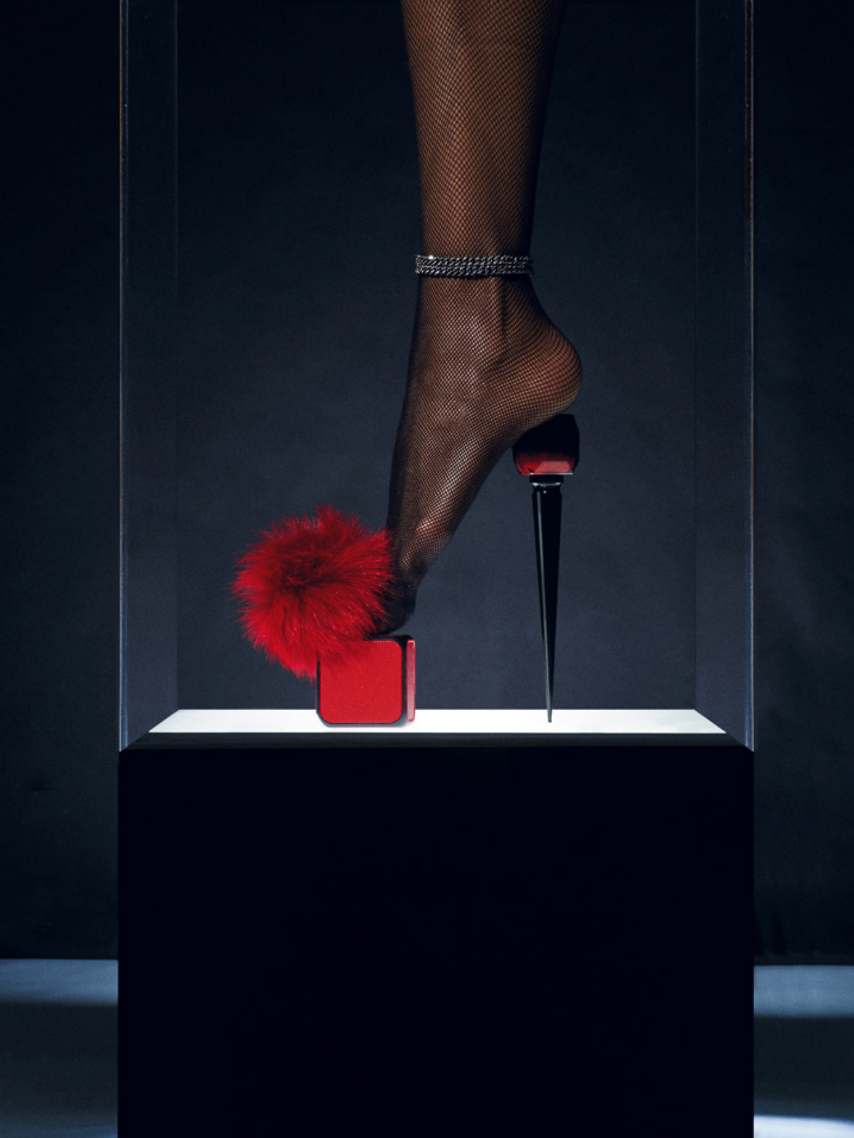 Close-up studio shot of a woman's leg wearing a black fishnet stocking and a red high heel with a fur pom-pom in a glass display case.