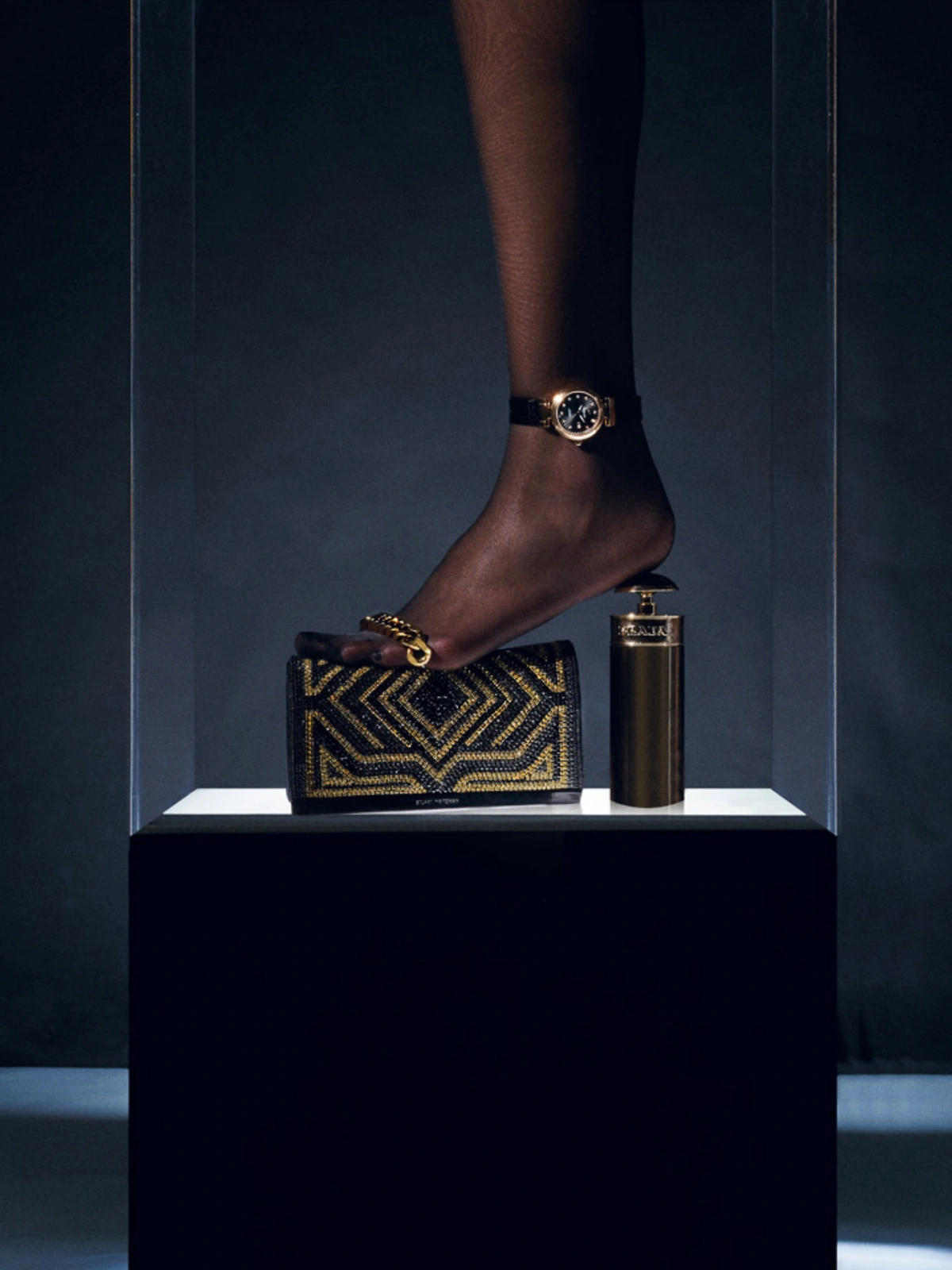 Close-up of a person's foot wearing a wristwatch as an anklet, stepping on a black and gold patterned wallet next to a cylindrical bottle.