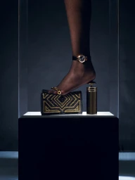 Close-up of a person's foot wearing a wristwatch as an anklet, stepping on a black and gold patterned wallet next to a cylindrical bottle.