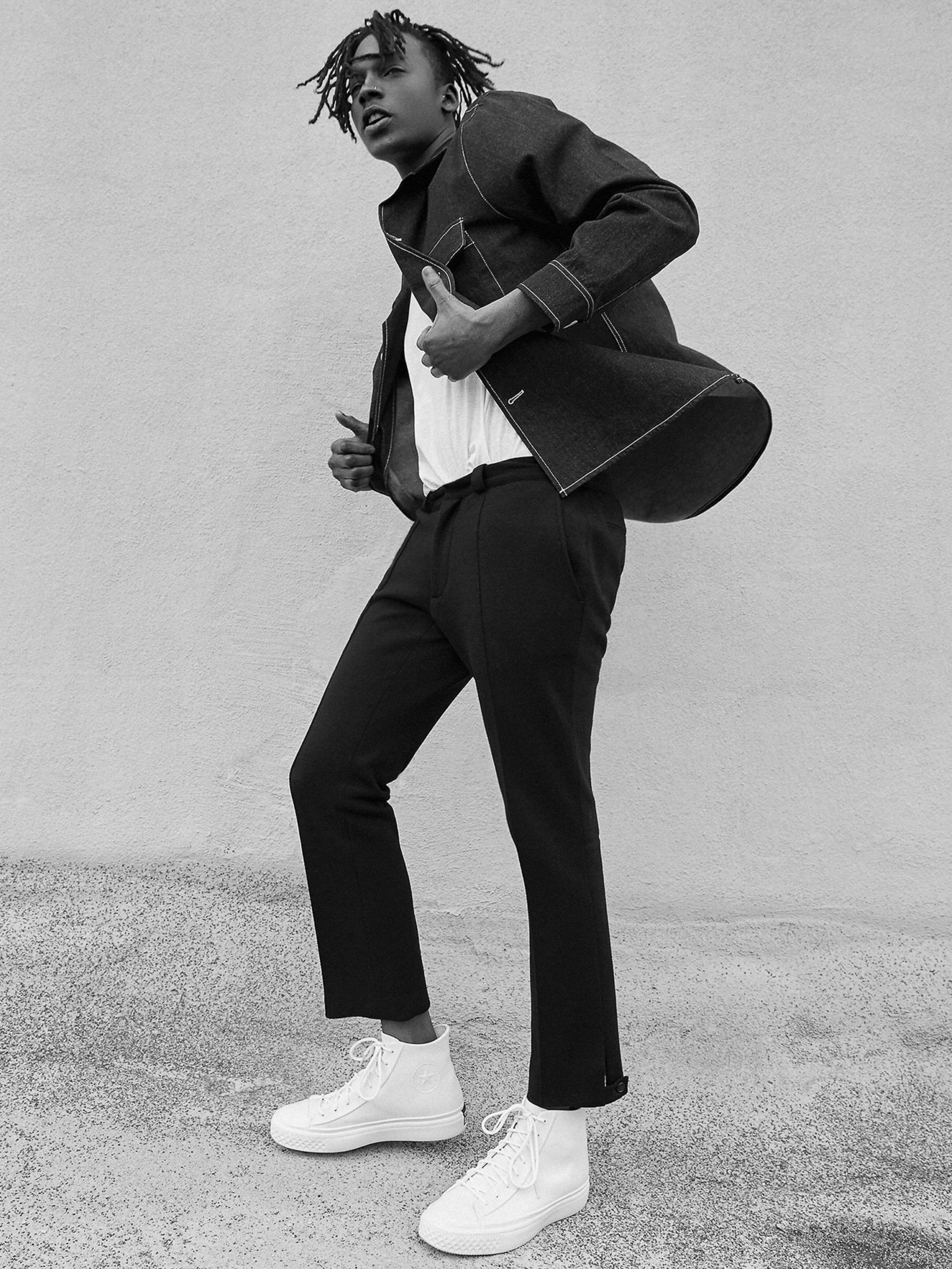 Stylish African American man in a denim jacket and white sneakers poses outdoors in a fashion shot. He looks at the camera.