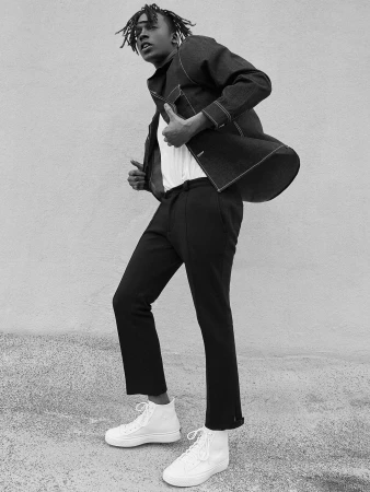 Stylish African American man in a denim jacket and white sneakers poses outdoors in a fashion shot. He looks at the camera.