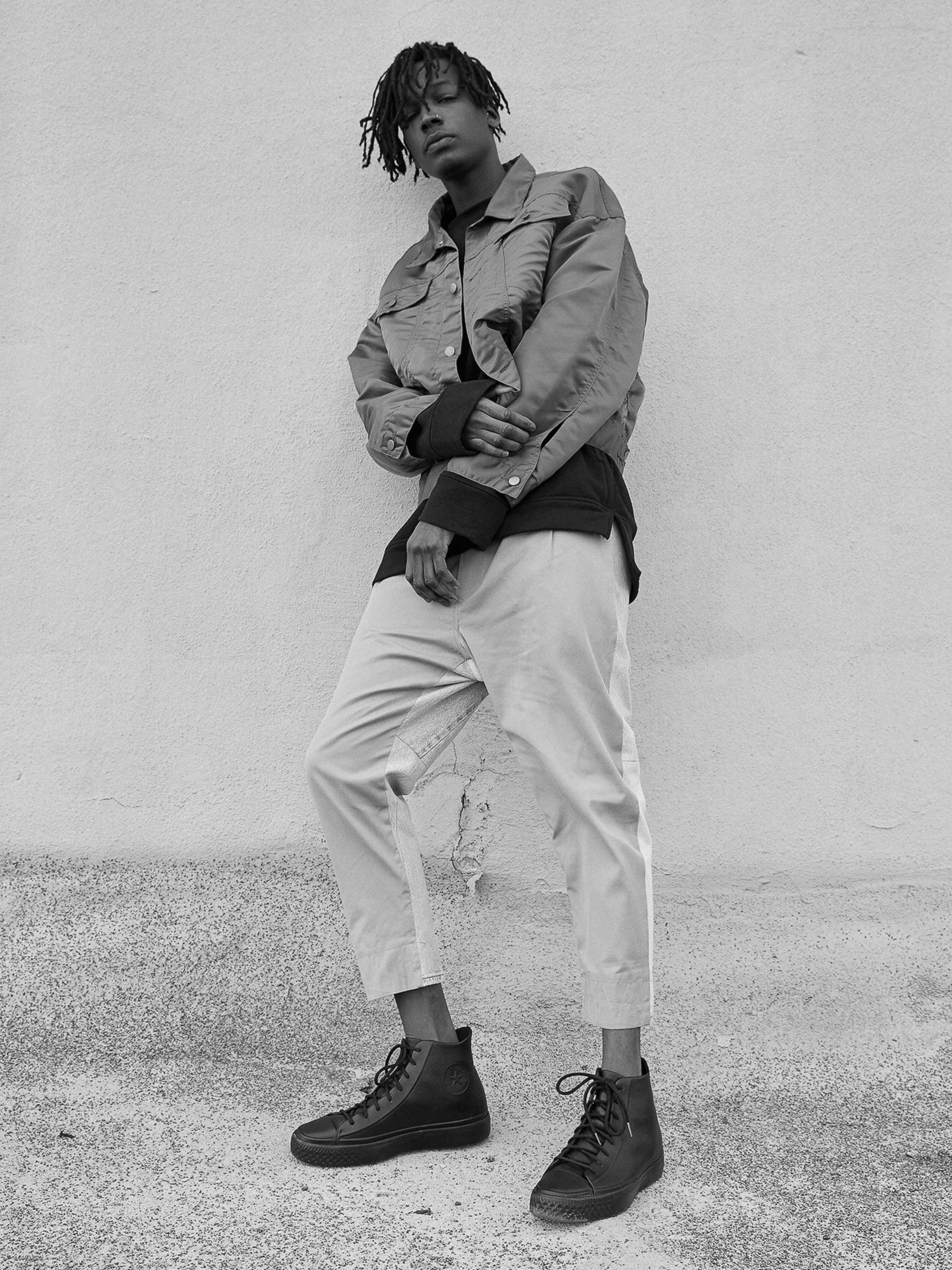 Young man with dreadlocks in casual jacket and pants poses against a textured wall, capturing urban style in black and white tones.