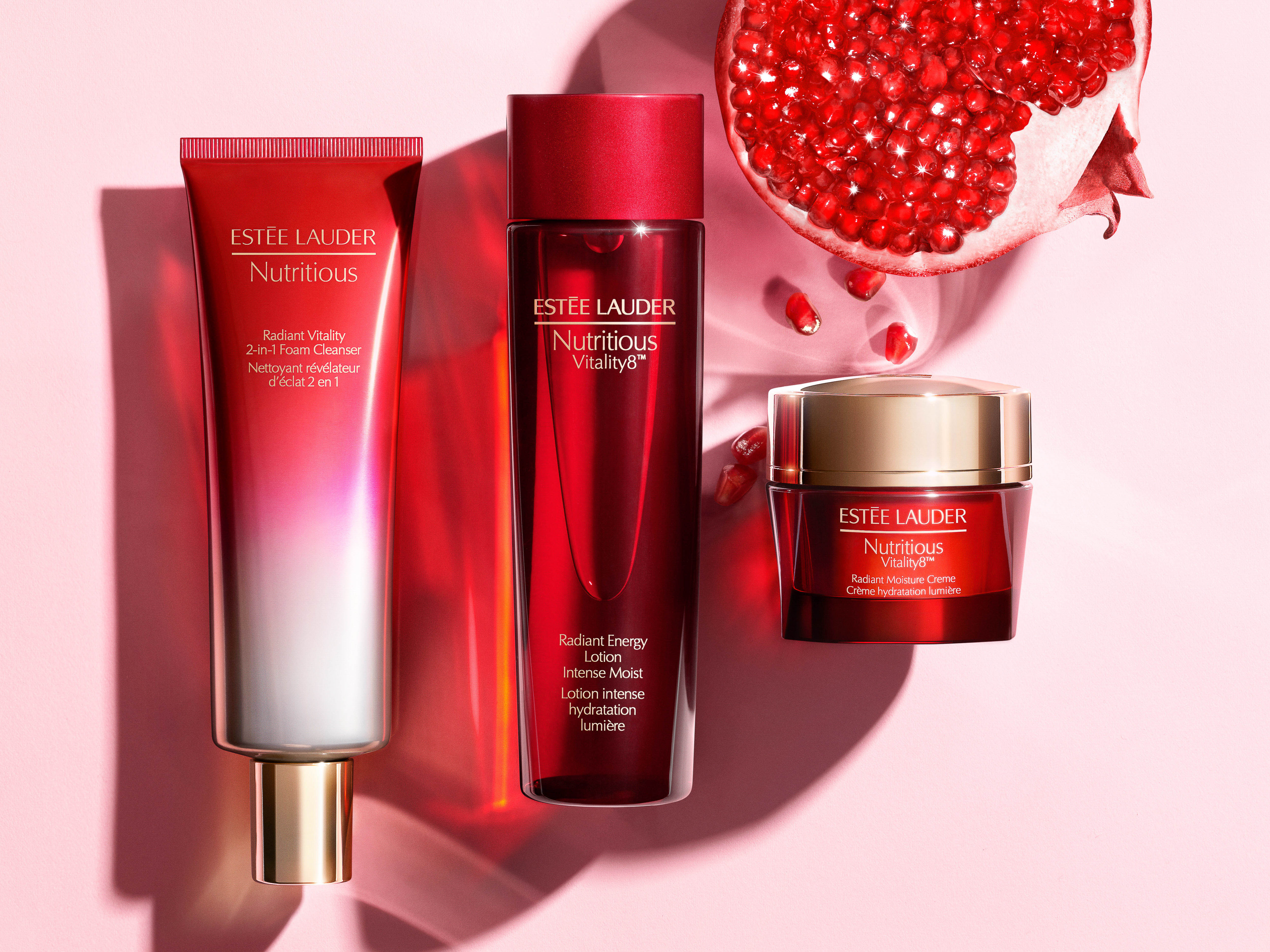 Three Estee Lauder Nutritious skincare products: face wash, lotion, and cream, are arranged on a light pink surface with a pomegranate half.