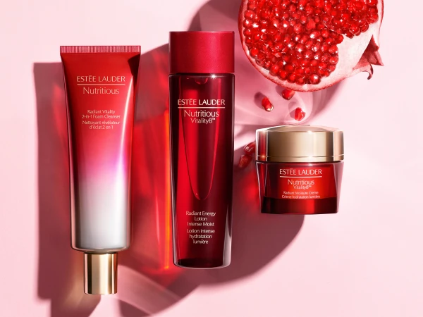 Three Estee Lauder Nutritious skincare products: face wash, lotion, and cream, are arranged on a light pink surface with a pomegranate half.