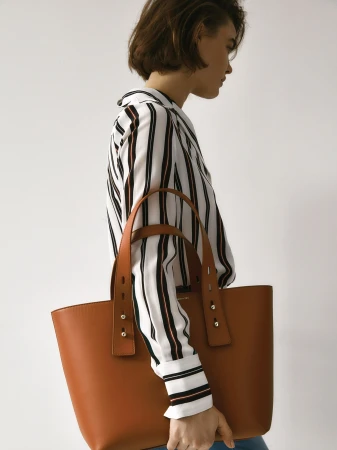 An adult woman stands in a photo studio holding a tan-colored leather tote bag and wearing a striped shirt against a white background.