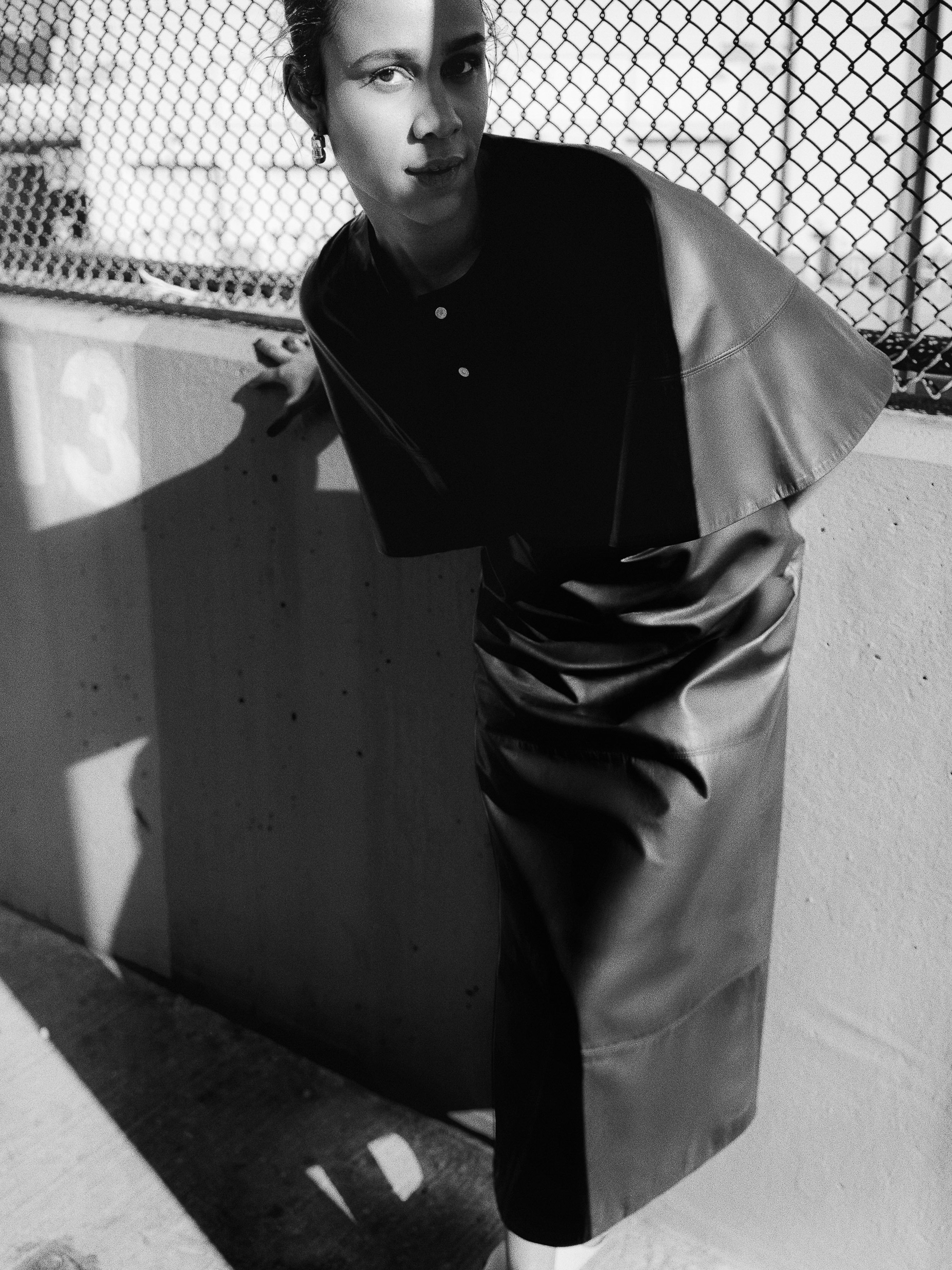 Black and white portrait of a woman wearing a cape and pencil skirt, leaning against a wall near a chain link fence.