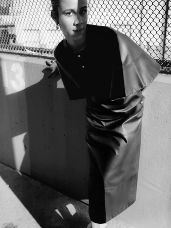 Black and white portrait of a woman wearing a cape and pencil skirt, leaning against a wall near a chain link fence.