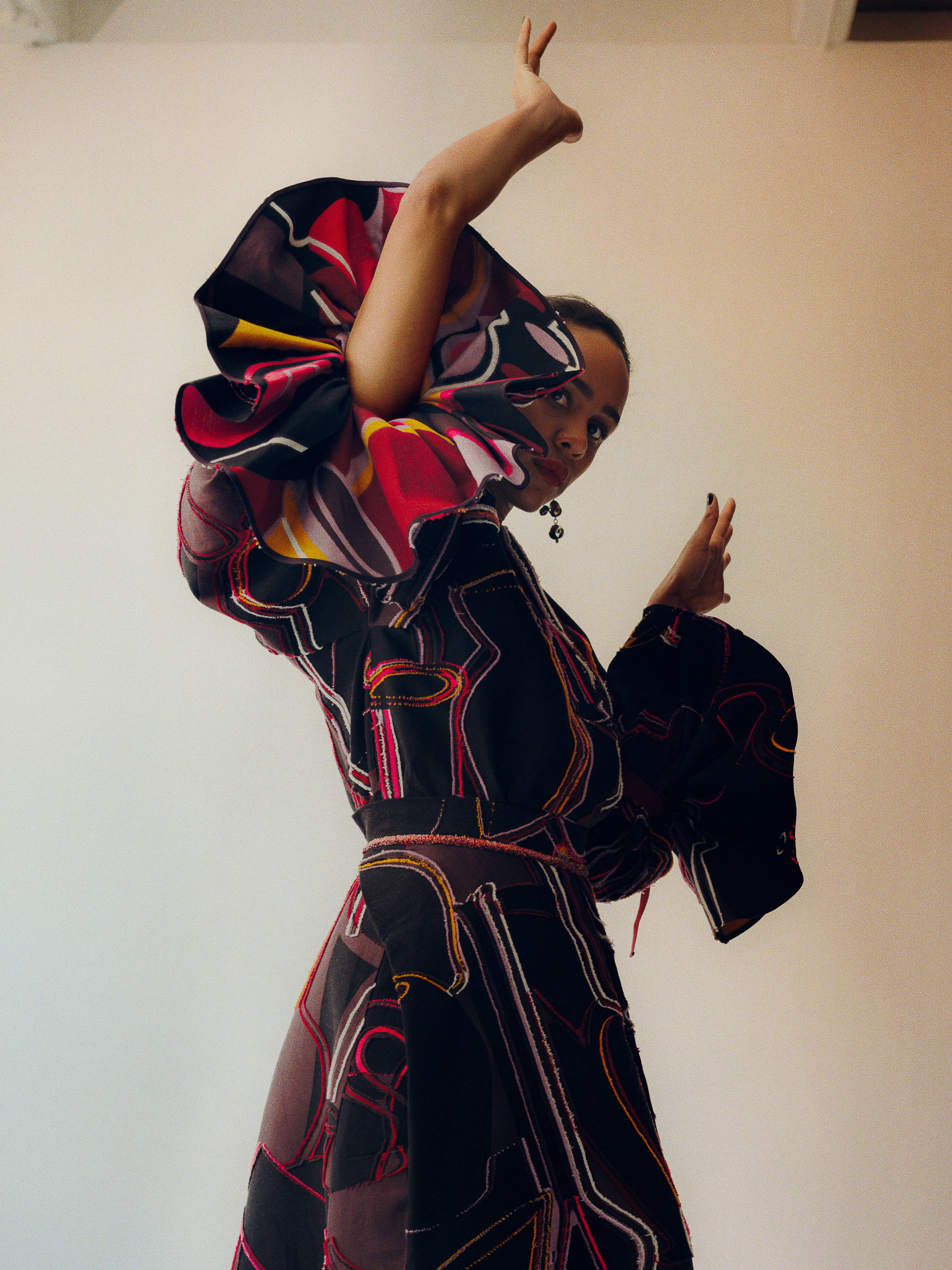 Portrait of a Black woman in a patterned dress with exaggerated sleeves striking a pose against a neutral wall, showcasing fashion.