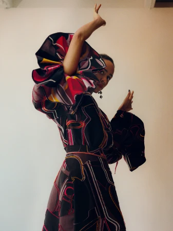 Portrait of a Black woman in a patterned dress with exaggerated sleeves striking a pose against a neutral wall, showcasing fashion.