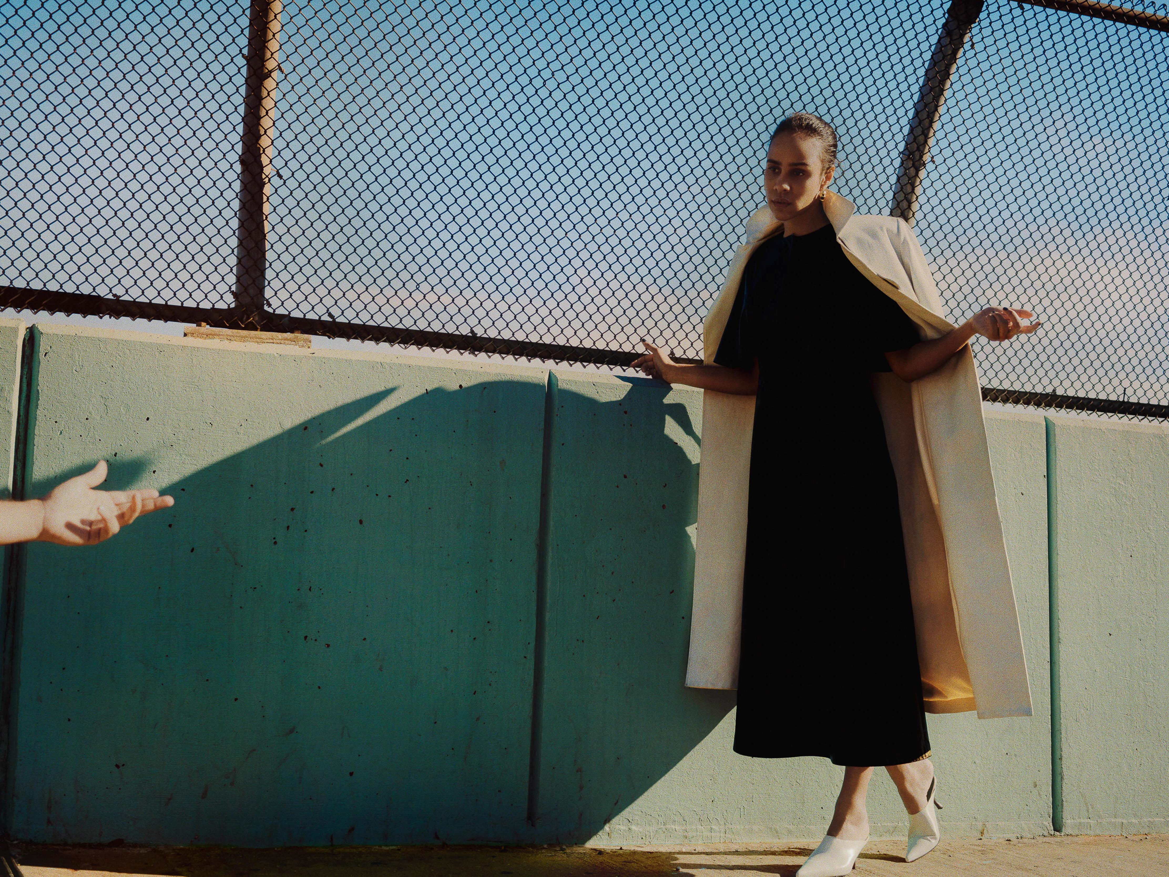 A woman in a black dress and light beige coat poses in front of a fence with an outstretched hand nearby, capturing a fashion moment.
