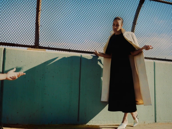 A woman in a black dress and light beige coat poses in front of a fence with an outstretched hand nearby, capturing a fashion moment.
