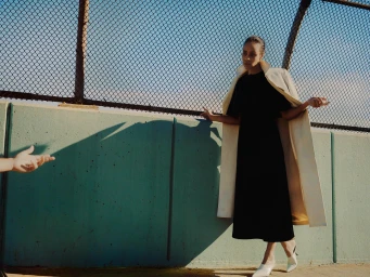 Woman in black dress and cream coat standing against a teal wall with a chain link fence above