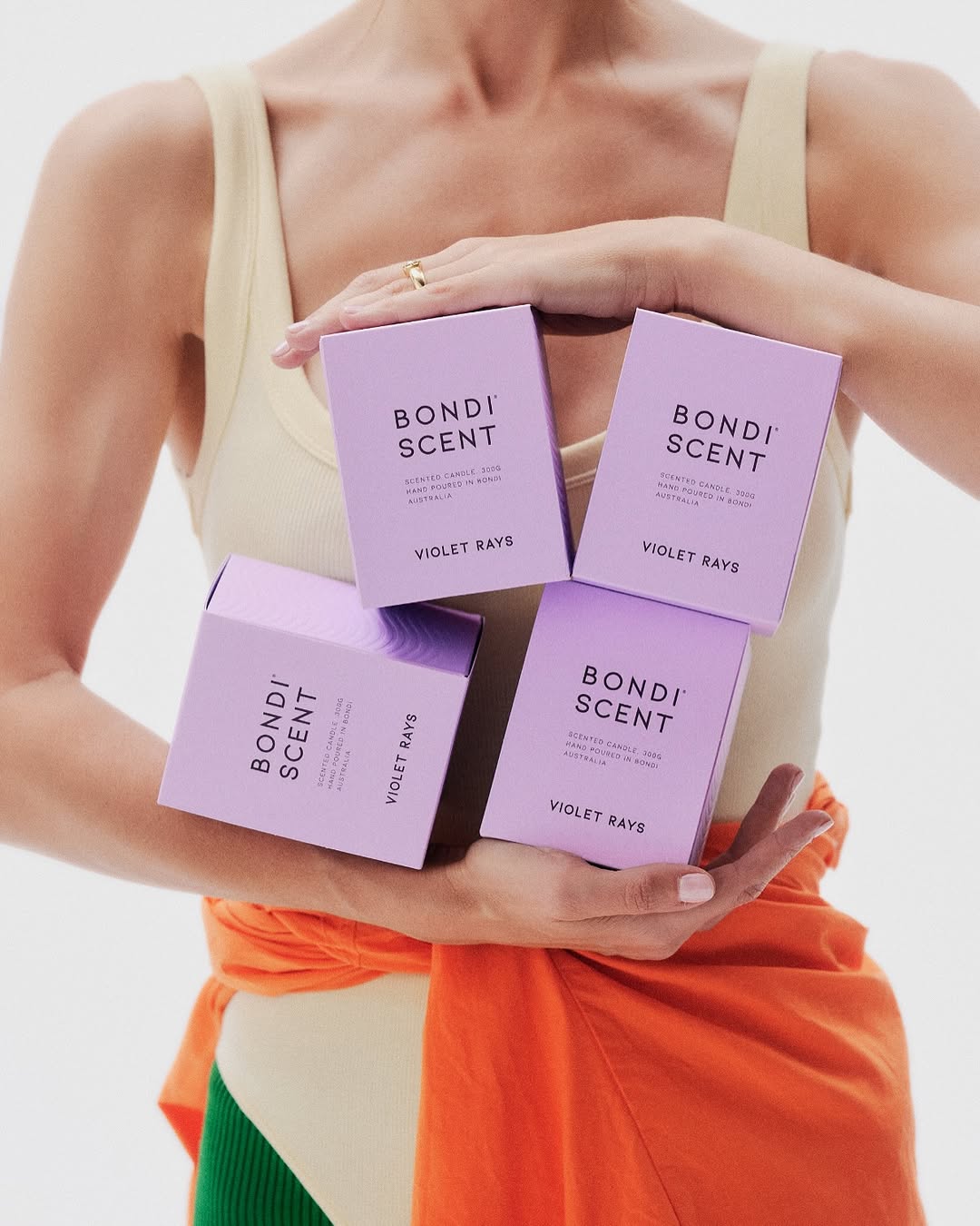 Close-up of an adult female holding four Bondi Scent Violet Rays candle boxes, set against a white backdrop and neutral tones.