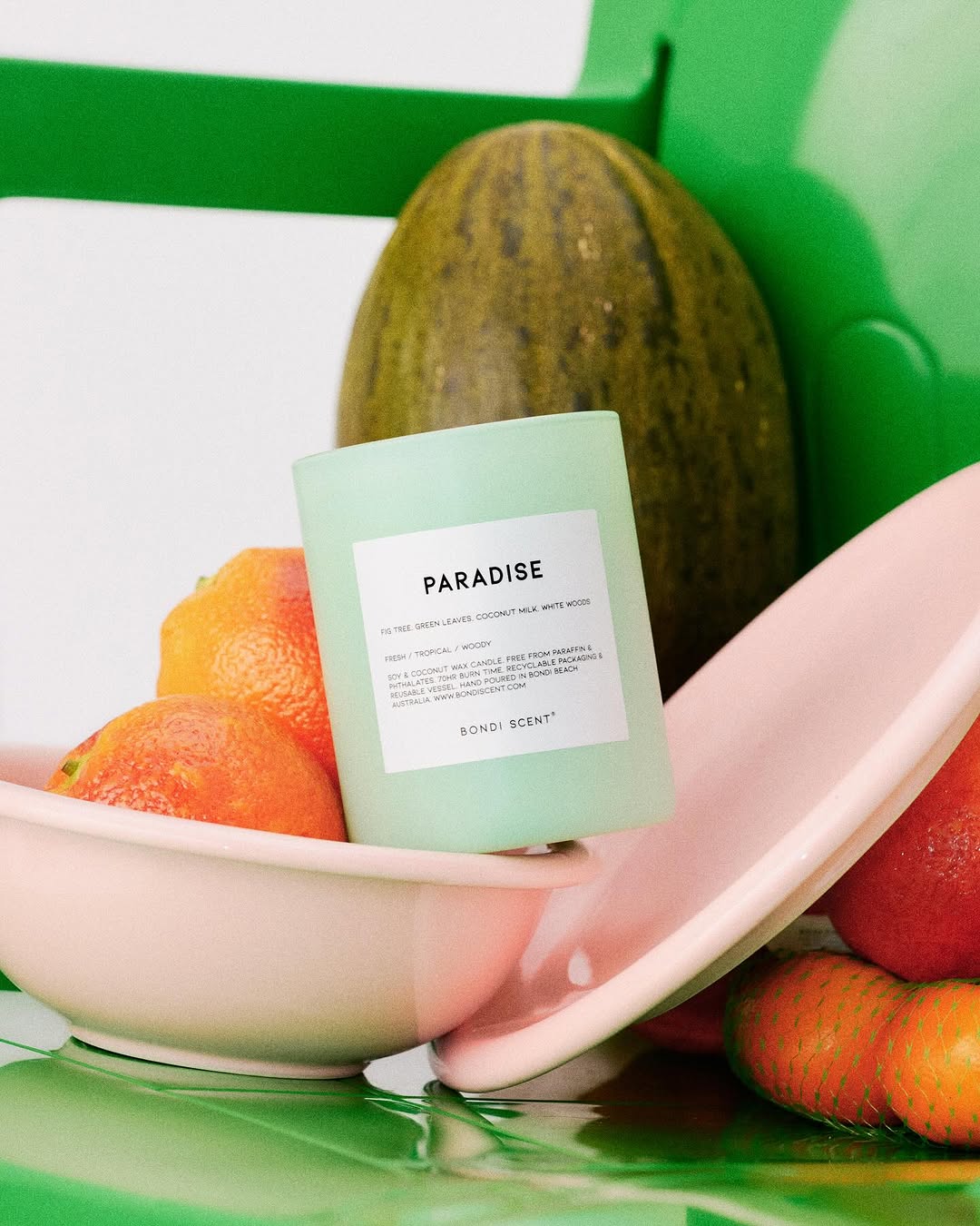 Close-up still life of a Bondi Scent candle, oranges, and melon on a green and pink reflective surface in a modern composition.