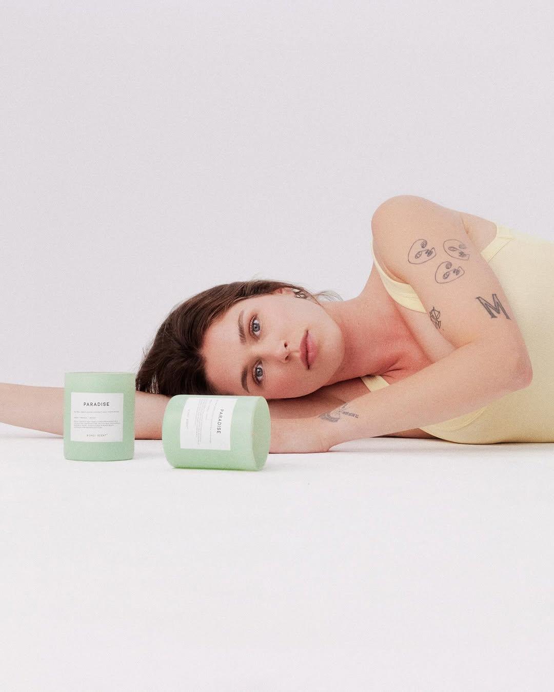 Woman in a yellow top poses on a white surface with light green cosmetic containers in a minimalist photo studio setting.