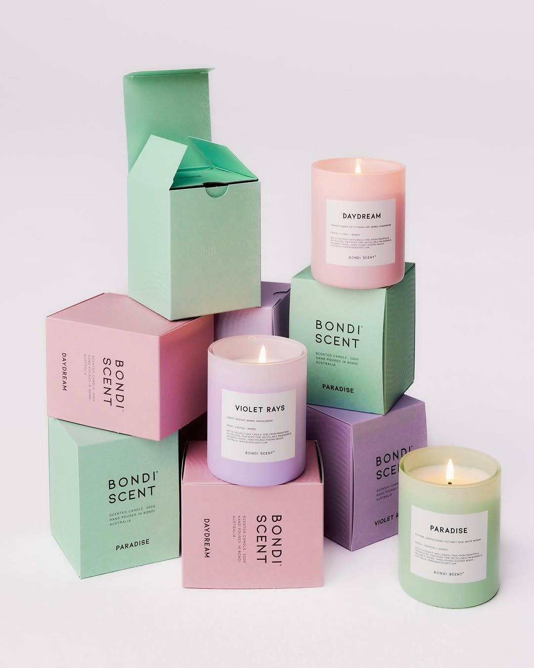 Several lit scented candles are displayed with their packaging boxes, featuring pastel shades like pink, purple, and mint green.