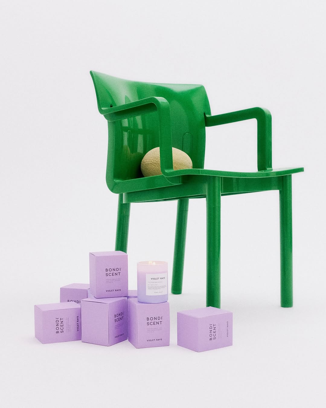 Green chair with ball, surrounded by lavender Bondi Scent candle boxes in a minimalist studio setting, showcasing product presentation.