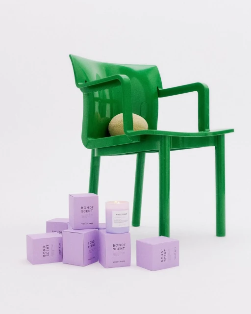 Green chair with ball, surrounded by lavender Bondi Scent candle boxes in a minimalist studio setting, showcasing product presentation.