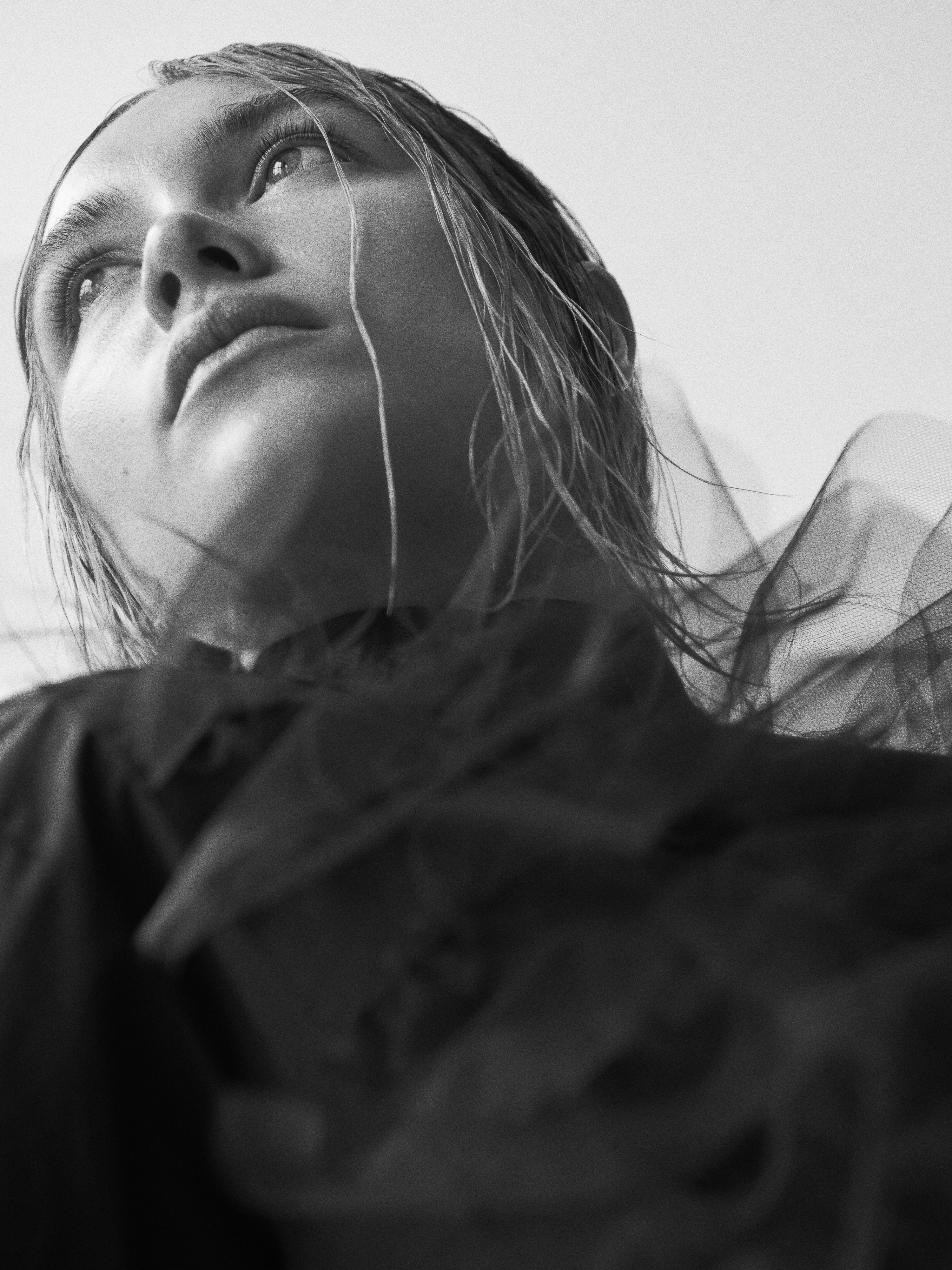 Black-and-white portrait of a young woman gazing upwards with light reflecting on her face, complemented by a sheer dark garment.