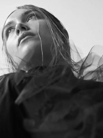 Black-and-white portrait of a young woman gazing upwards with light reflecting on her face, complemented by a sheer dark garment.