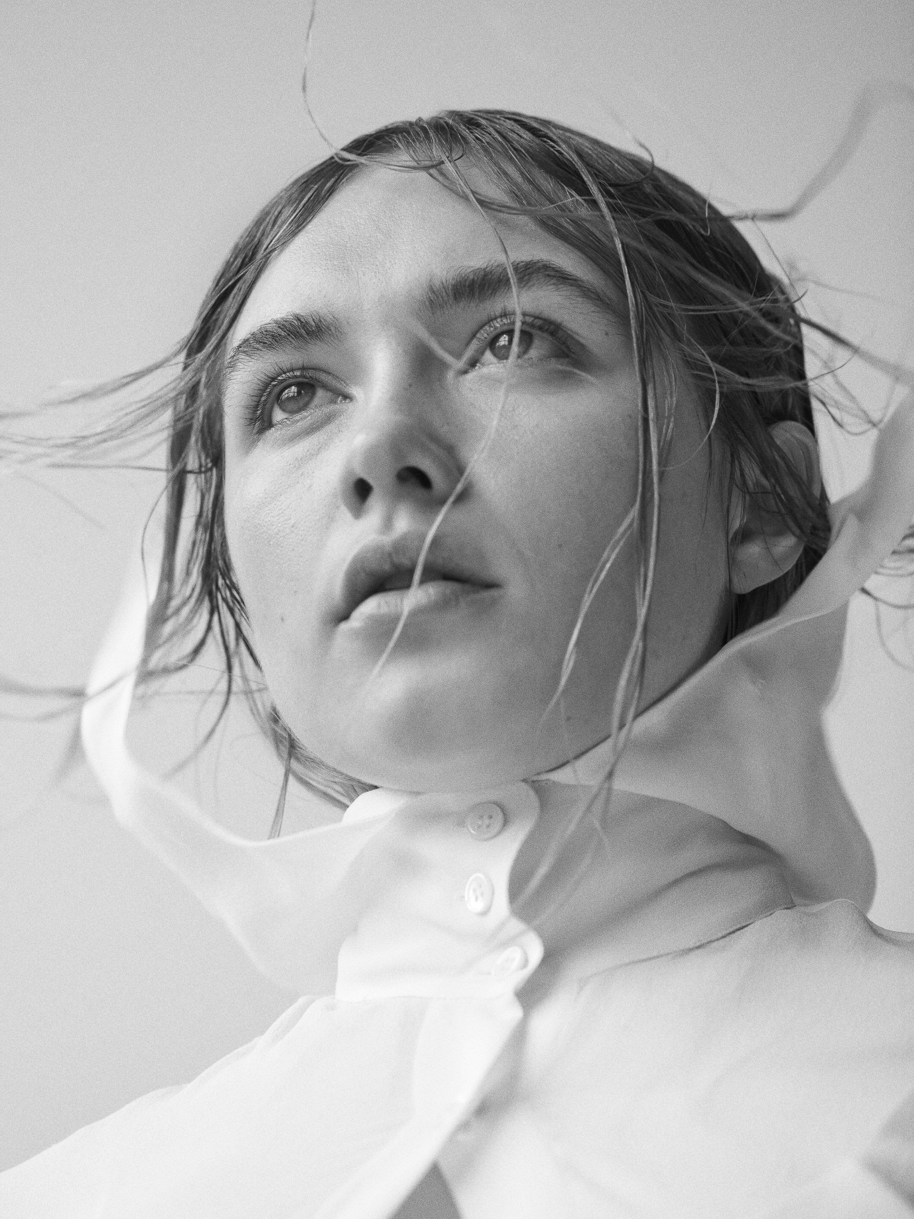 Monochrome portrait of a young woman with wet hair and a white collared shirt, looking upwards against a pale background in a studio.
