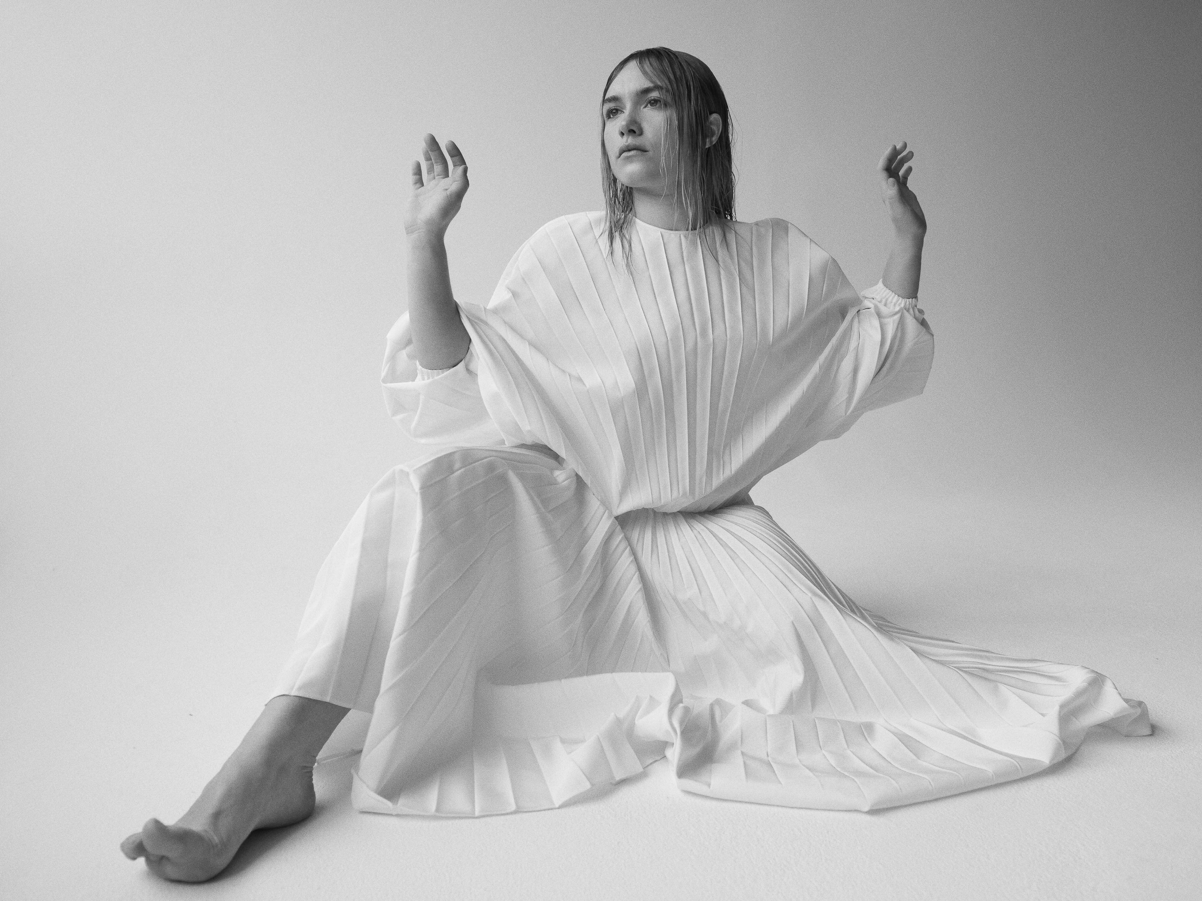 Monochrome shot of a woman in a pleated white dress, seated with raised hands, against a pale background, creating a minimalist and ethereal feel.