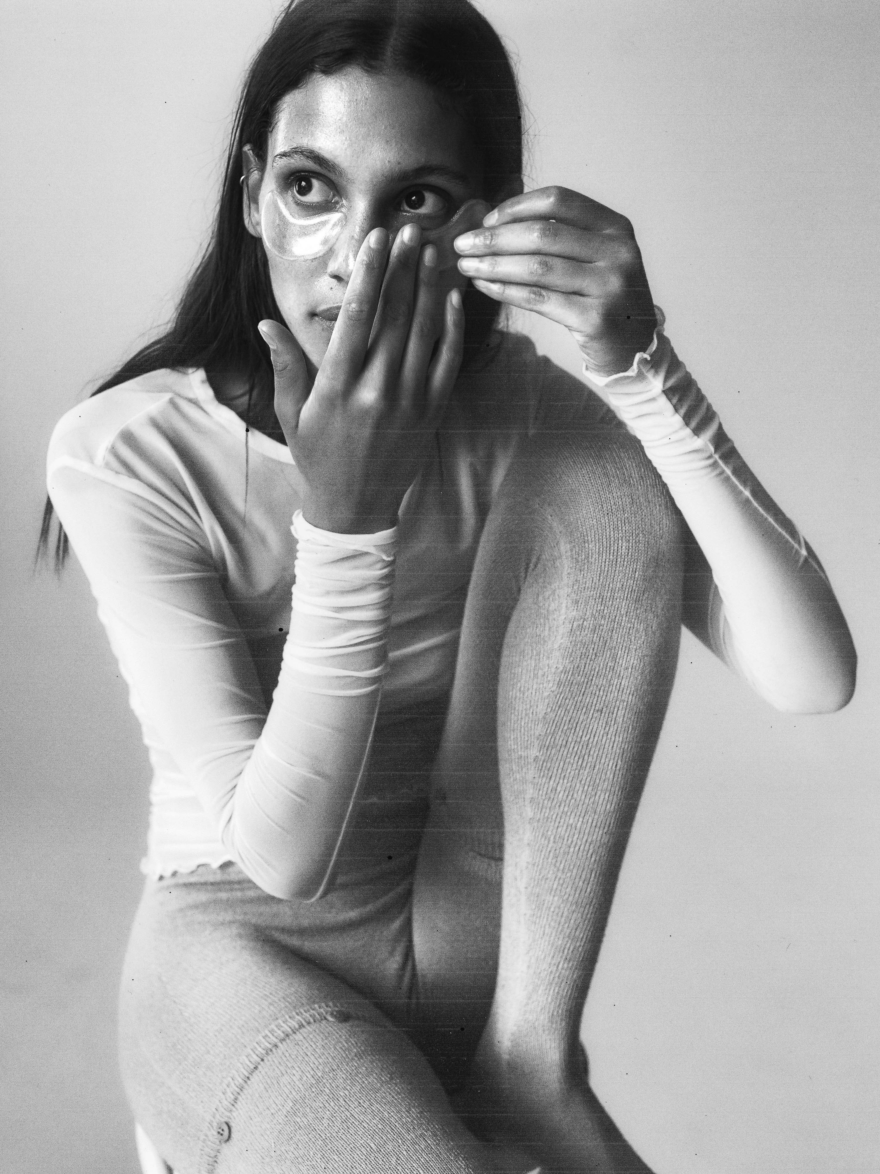 A woman with long, dark hair applies eye mask patches in a black and white, medium shot, wearing a sheer top, showcasing a beauty routine.