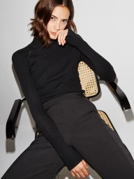 Model wearing a black ribbed turtleneck sweater and dark pants seated on a cane-backed chair against a light wall.