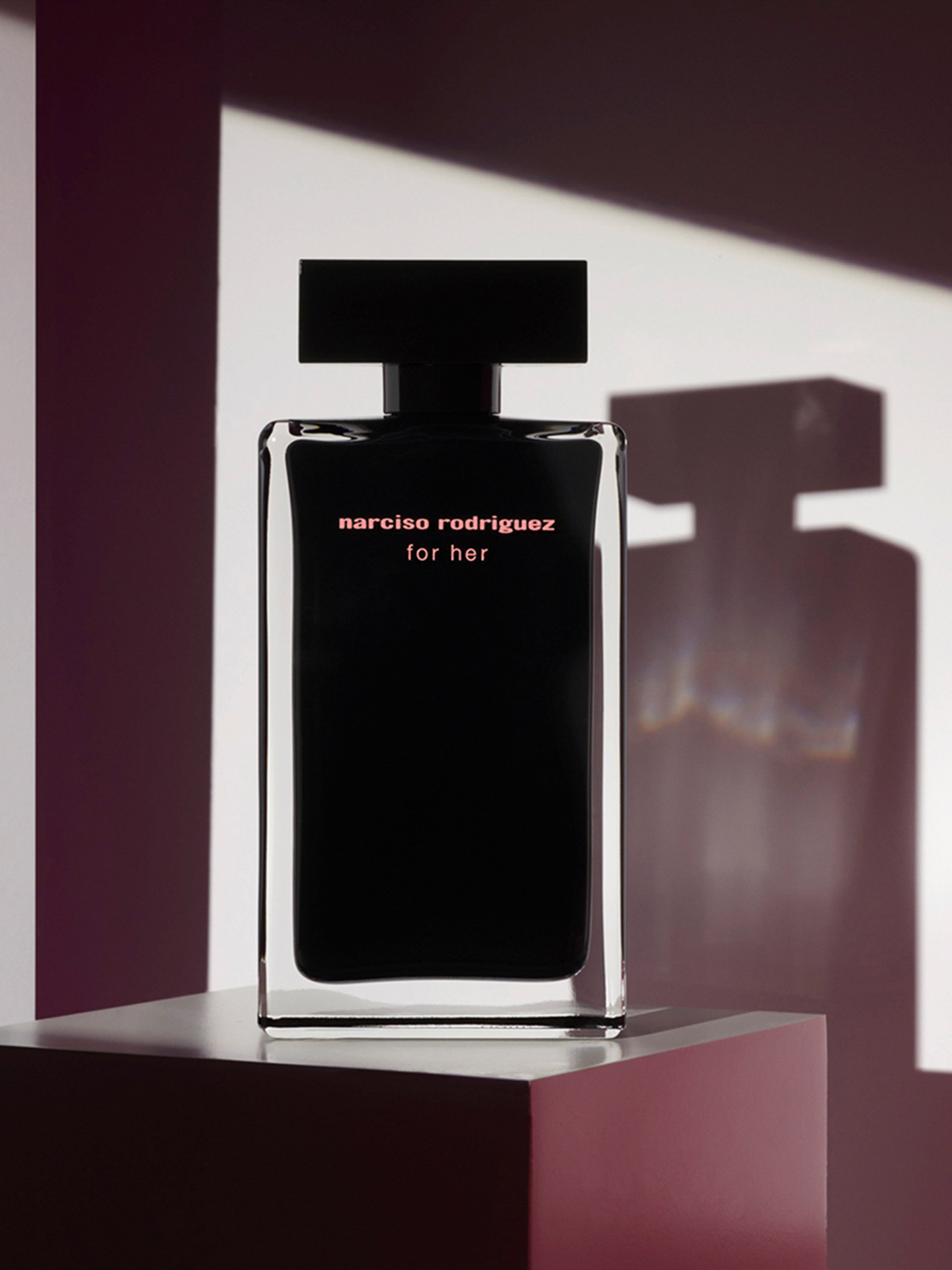 A black Narciso Rodriguez perfume bottle stands on a plinth with geometric shadows against a white backdrop in a studio shot.