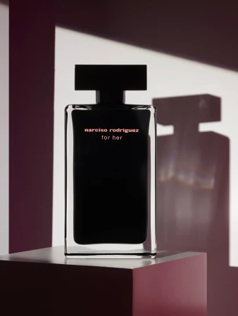 A black Narciso Rodriguez perfume bottle stands on a plinth with geometric shadows against a white backdrop in a studio shot.