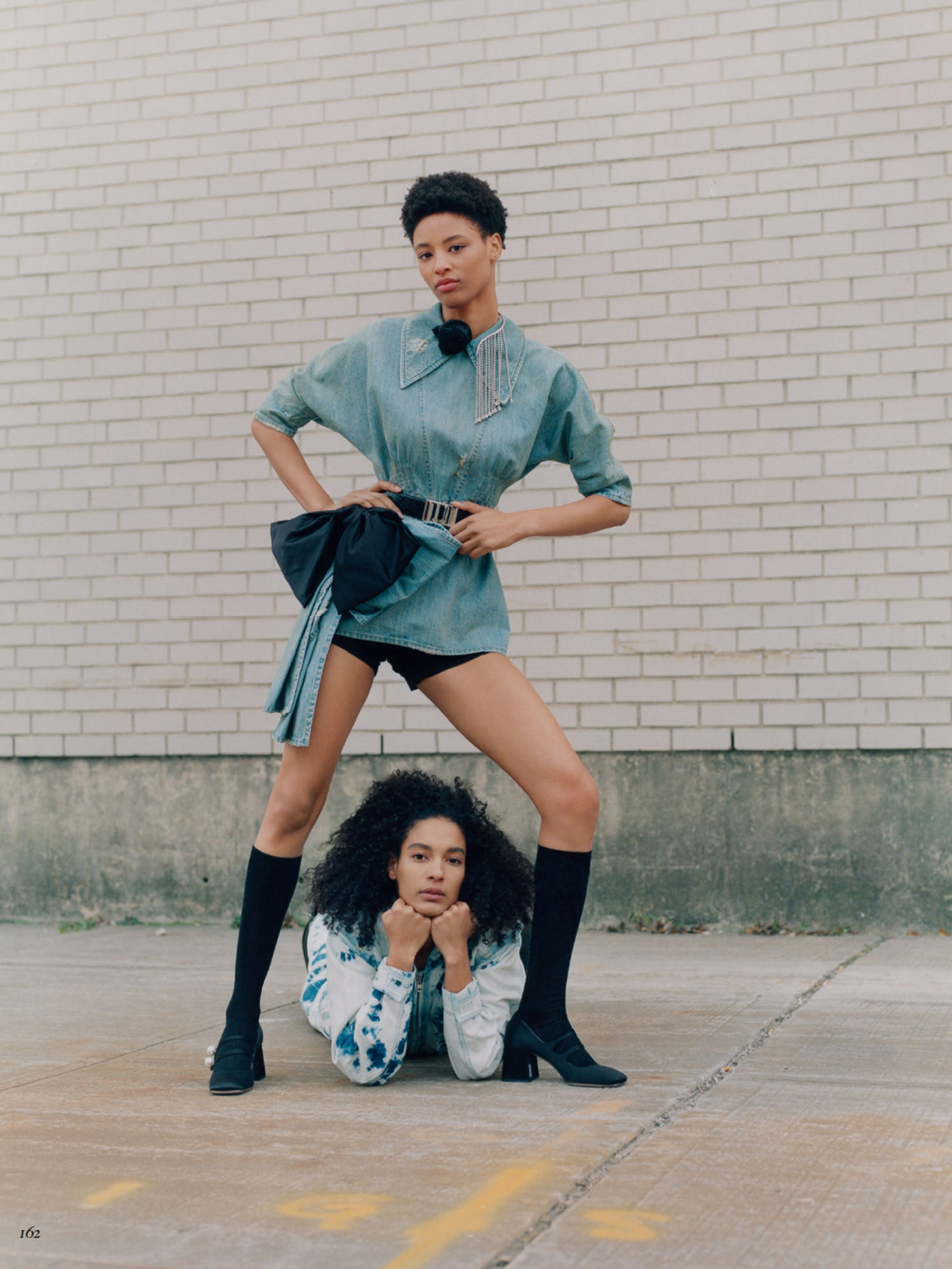 Two women posing outdoors in a fashion editorial. One stands wearing a blue shirt dress and the other lies on the ground.