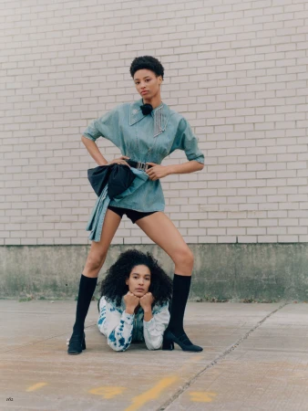 Two women posing outdoors in a fashion editorial. One stands wearing a blue shirt dress and the other lies on the ground.