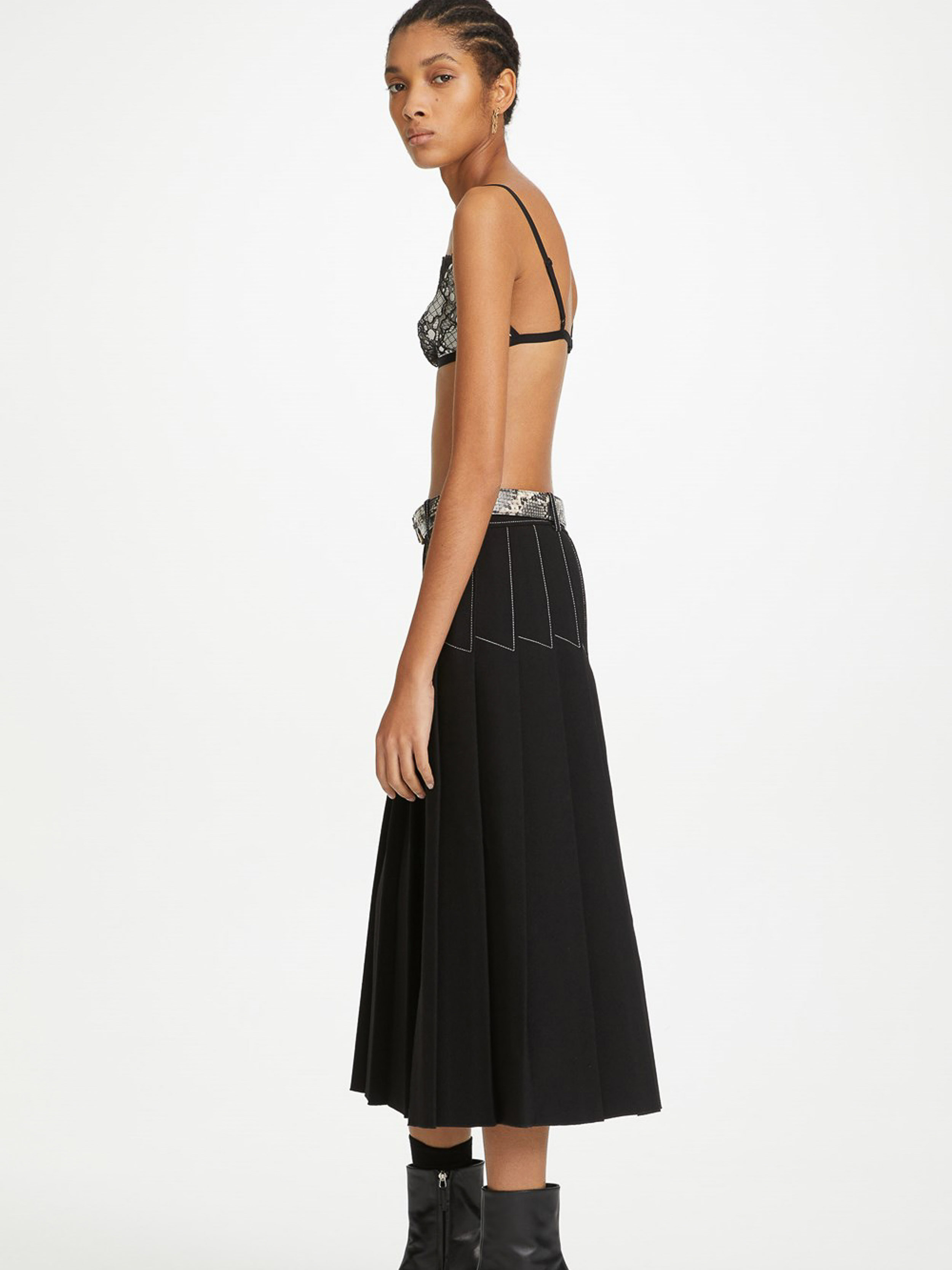 Side profile of a woman in a black bralette, black skirt with white stitching, belt, and boots, standing in a white photo studio.