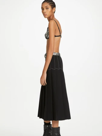 Side profile of a woman in a black bralette, black skirt with white stitching, belt, and boots, standing in a white photo studio.