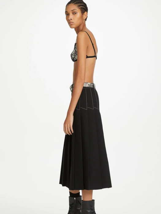 Model in black pleated skirt and patterned top standing against a plain white background for Dion Lee.