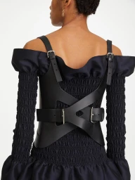 Rear view of a model wearing a black leather harness over a dark off-the-shoulder dress against a white background
