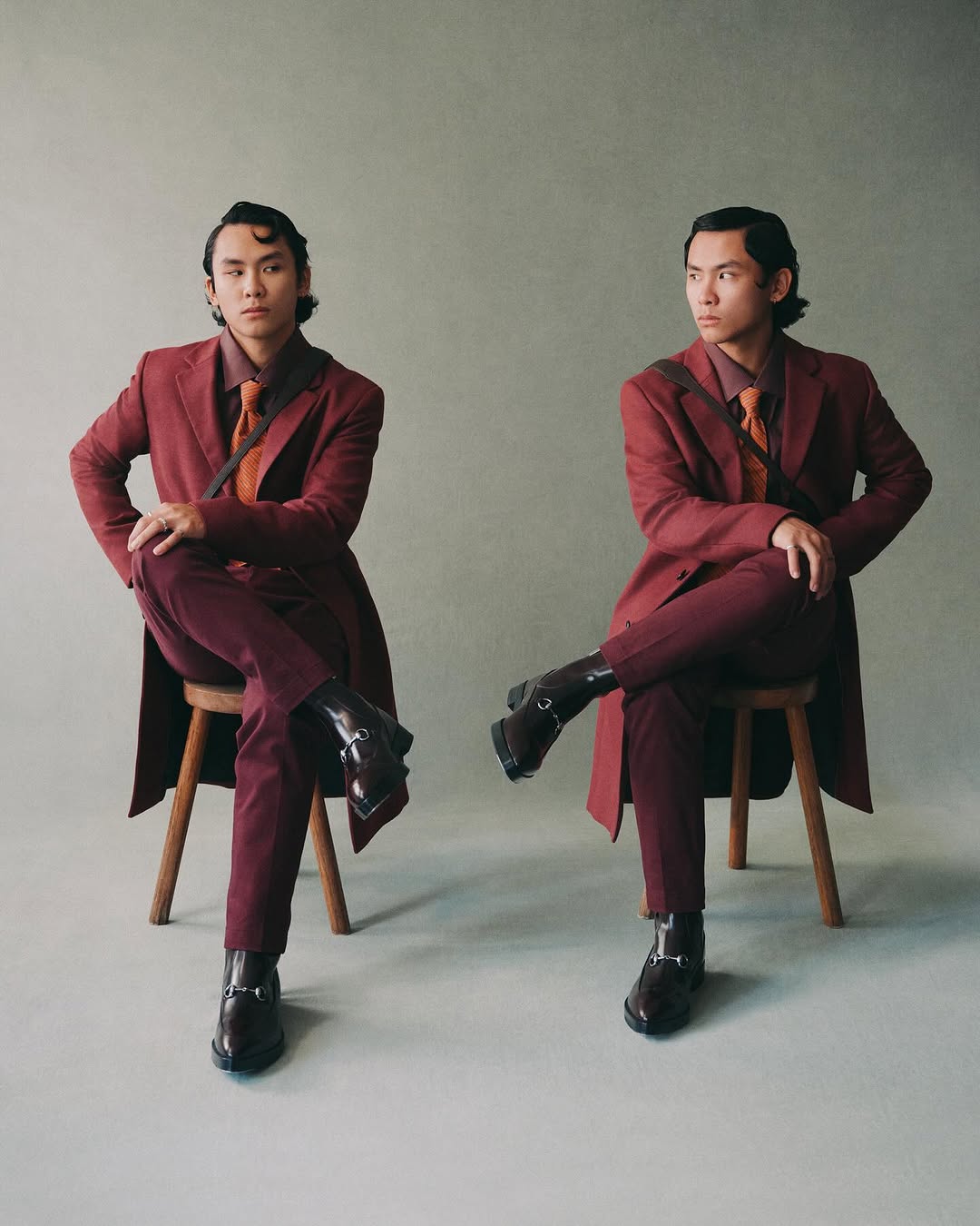 Two men in matching burgundy suits pose sitting on wooden stools in a studio setting, facing opposite directions, showcasing fashion.