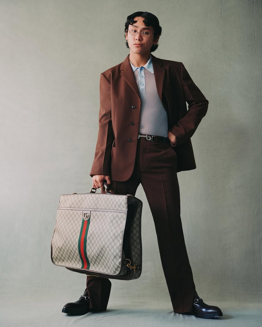 An Asian male model poses in a brown suit with a patterned briefcase, wearing a light blue polo shirt and shiny black dress shoes in studio.