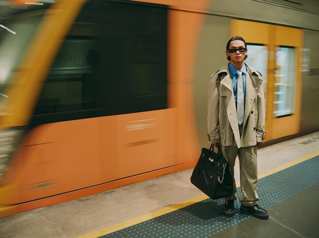 A person in a tan trench coat and sunglasses stands on a train platform with a black bag, as an orange train passes in the background.