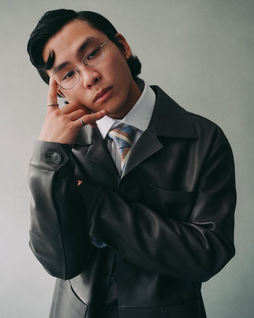 Portrait of an Asian man wearing glasses and a suit, with his hand near his face, against a neutral backdrop. Fashion and style.