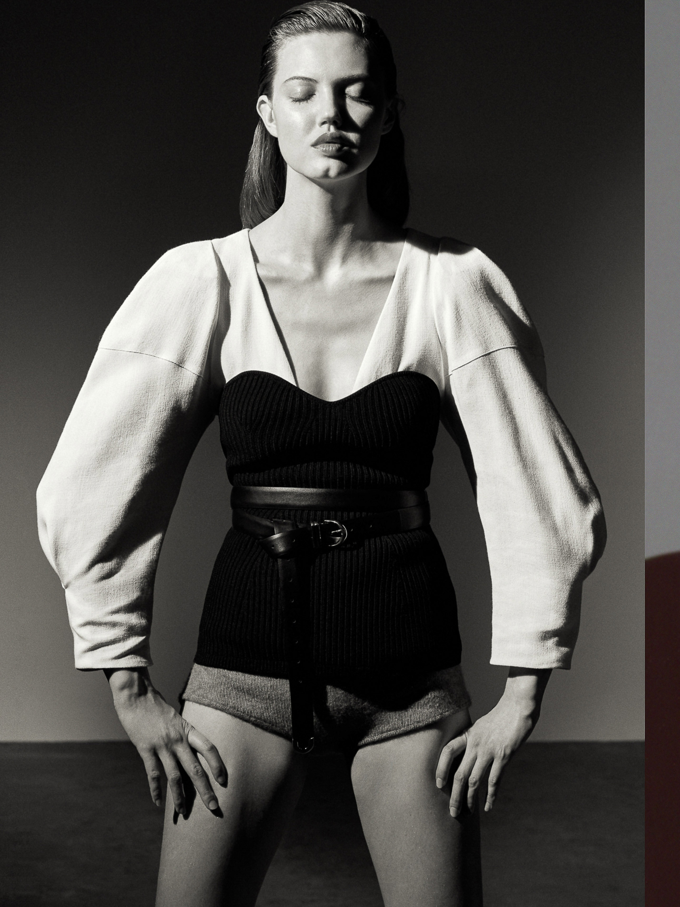 A model poses indoors wearing a black knit top with a belt, puffy-sleeved off-white jacket, and shorts in a studio setting with soft lighting.