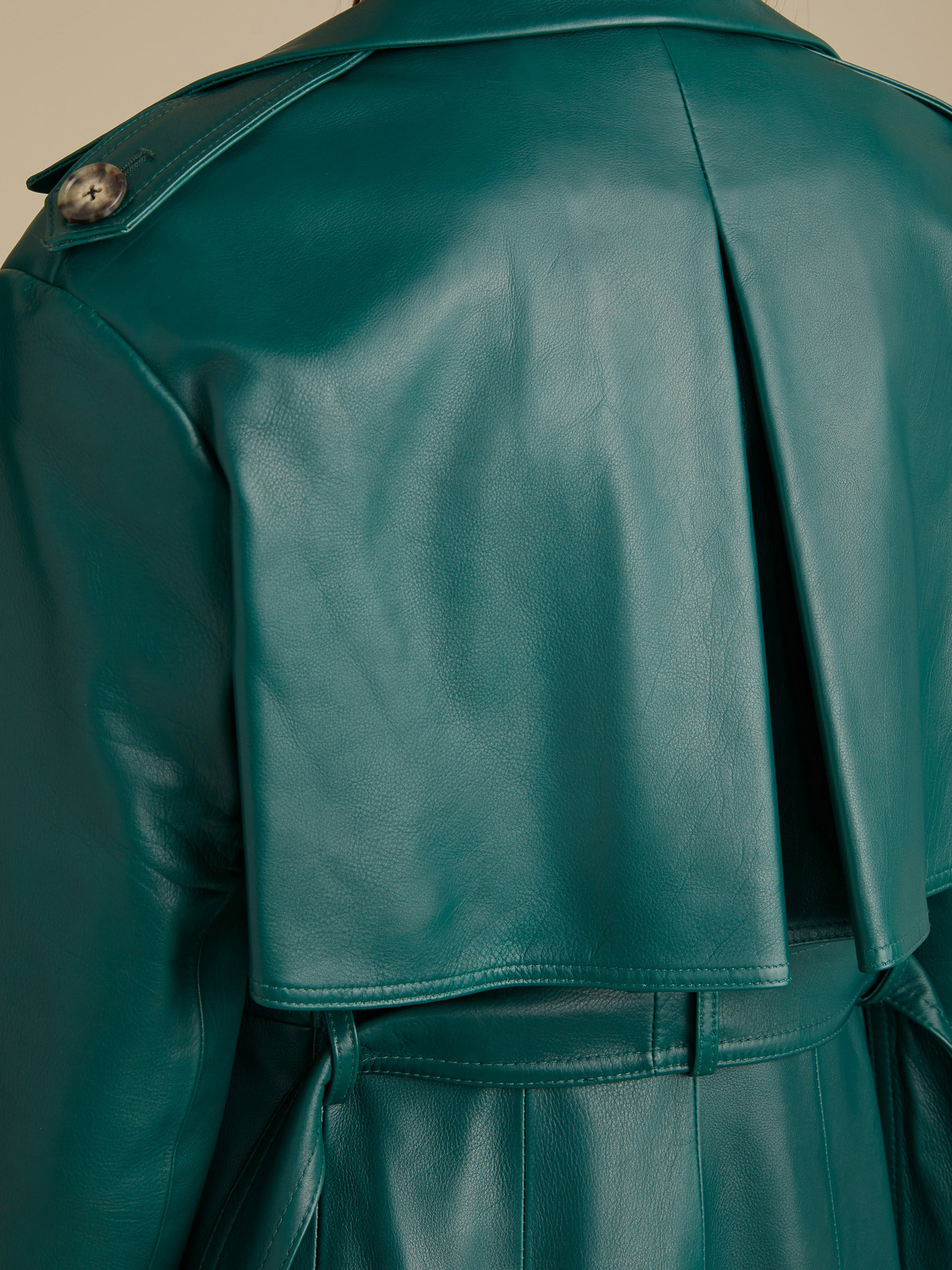 Close-up of a person wearing a teal, belted leather trench coat. The photo focuses on the back of the coat, including a buttoned epaulette.