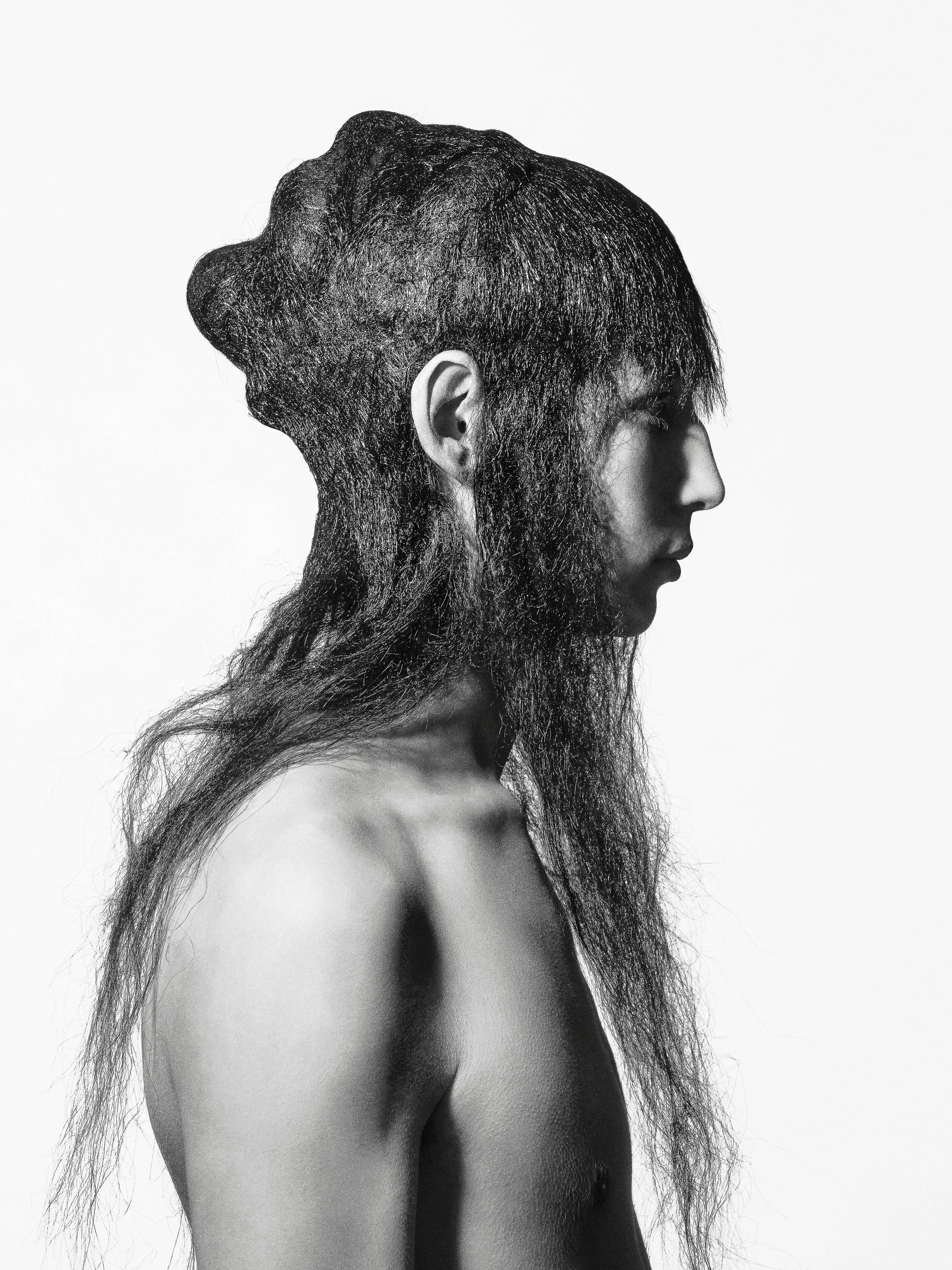 Monochrome profile of an adult with long, dark hair covering the head and chest, against a plain white backdrop in a studio setting.