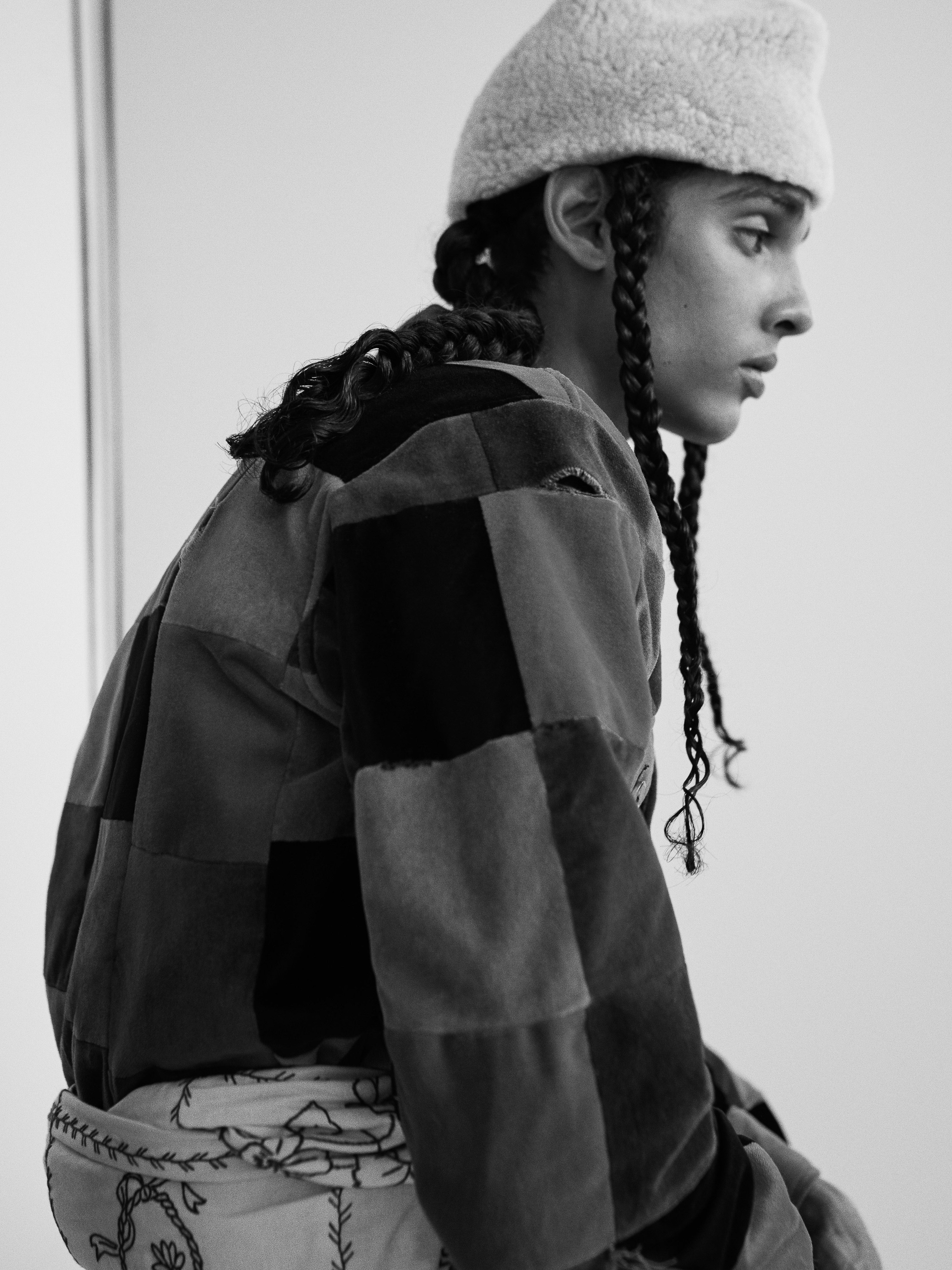 A portrait of a person with braids wearing a shearling hat and patchwork jacket against a neutral background, black and white aesthetic.