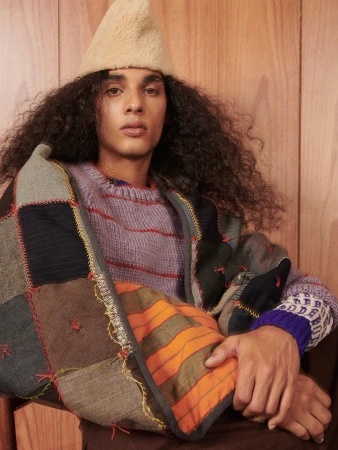 Man with long curly hair wears a patchwork jacket, knit sweater, and a knit cap, posing indoors in front of a wood wall.