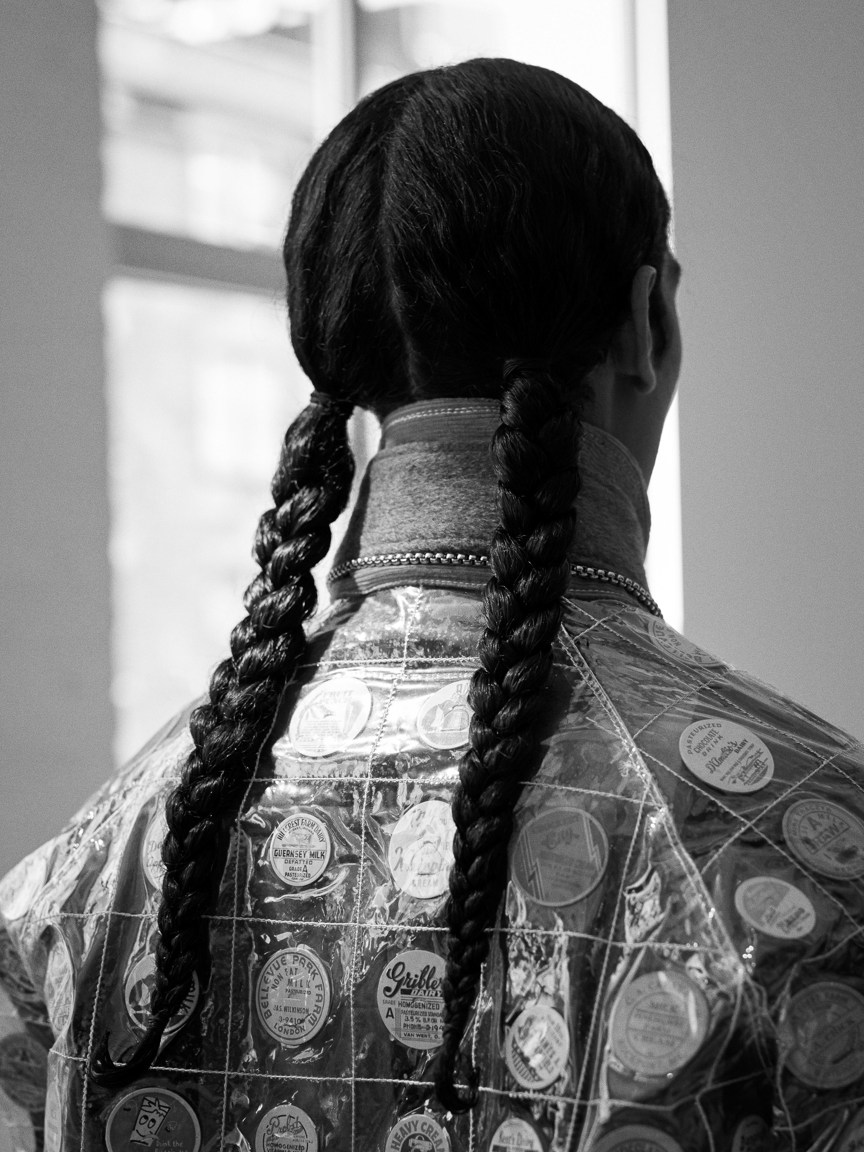 A person with braided hair, from the rear, wearing a clear plastic jacket with round decorations, stands in front of a window in black and white.