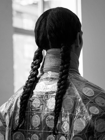 A person with braided hair, from the rear, wearing a clear plastic jacket with round decorations, stands in front of a window in black and white.
