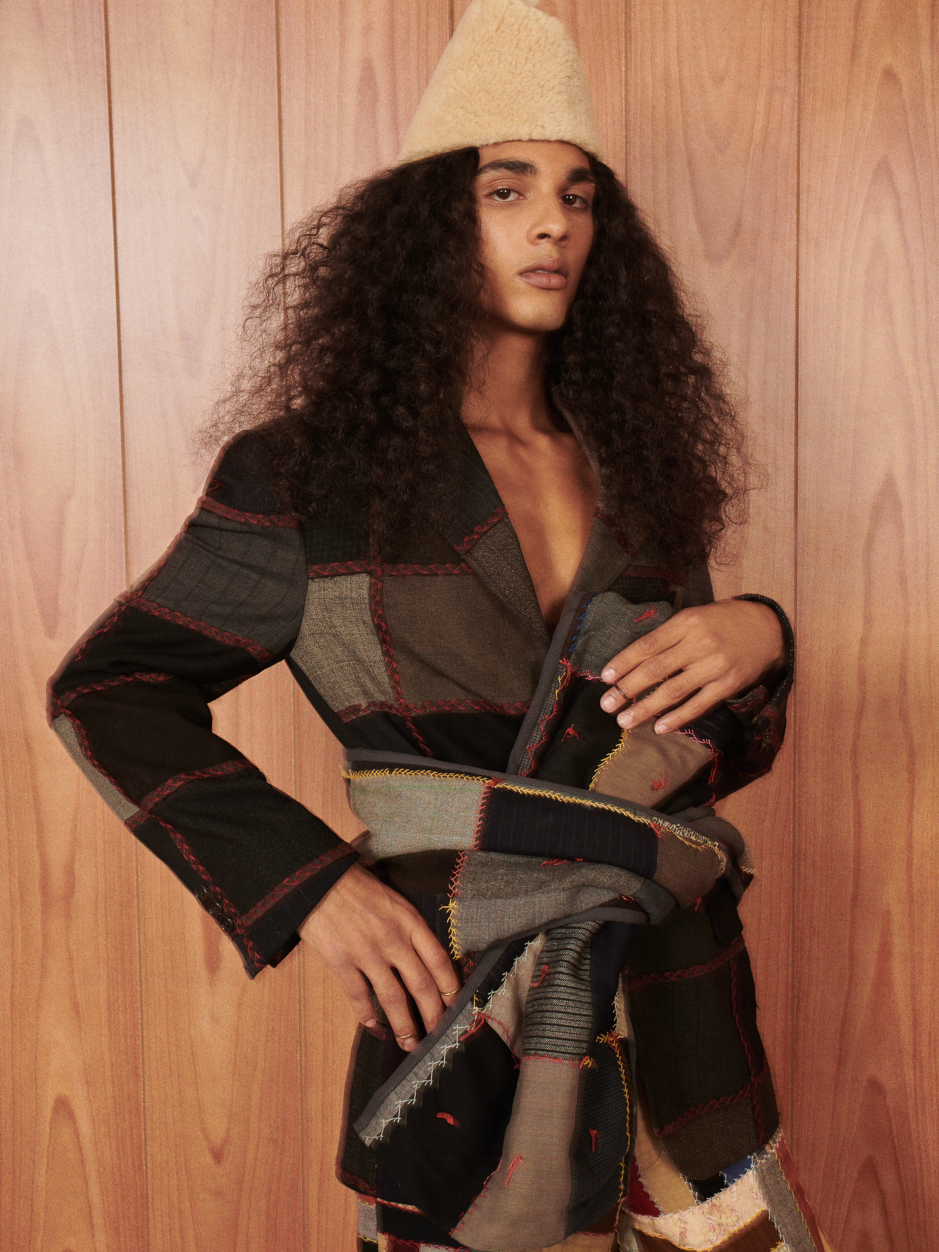 Portrait of a young adult with long curly hair wearing a tan cone hat and patchwork blazer against a wood panel background.