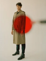 Young man in a beige trench coat standing against a pale background with a blurred red graphic element overlay.