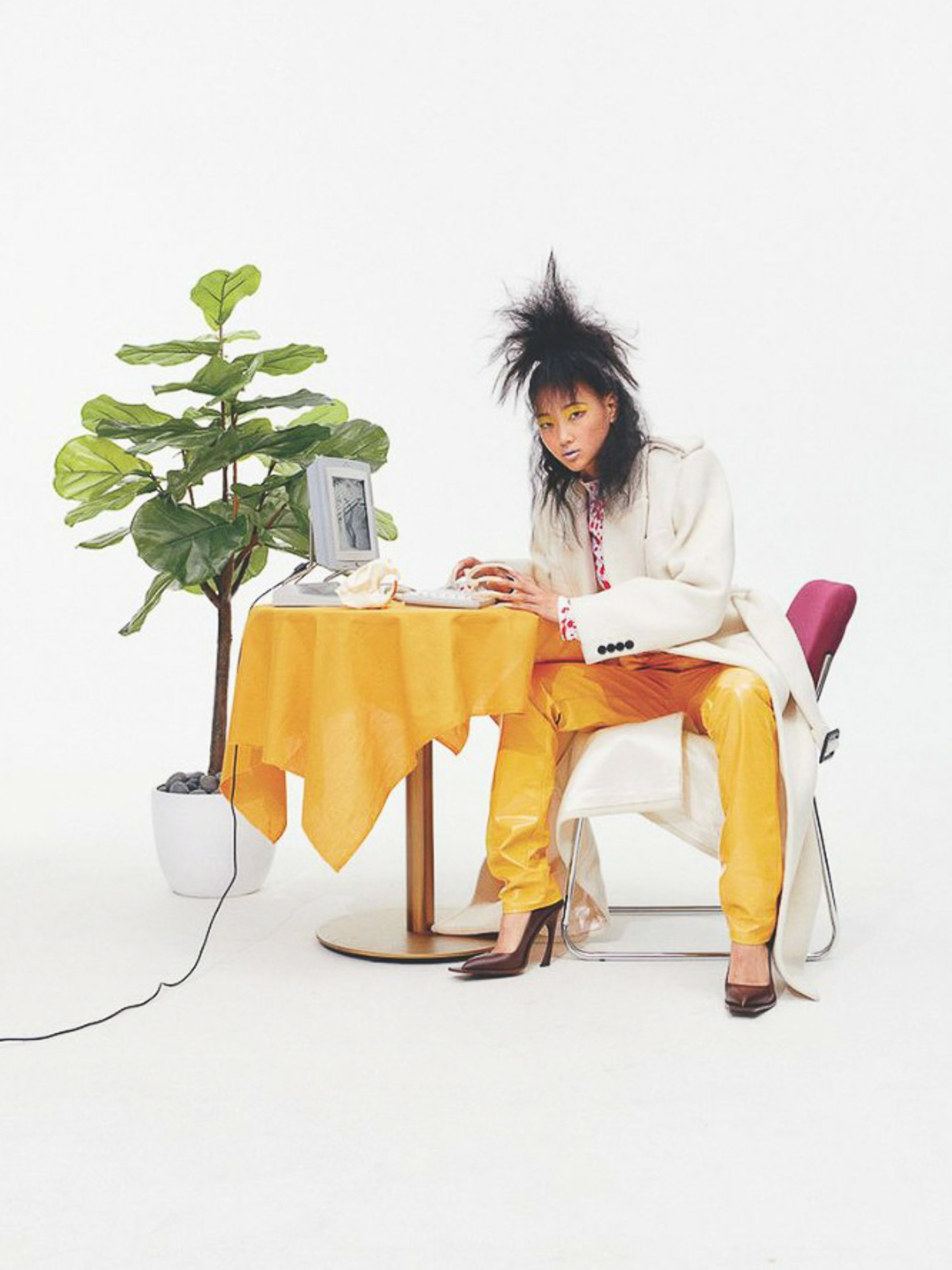 Woman with yellow pants poses at a table with a retro computer and potted plant against a white background in a studio setting.